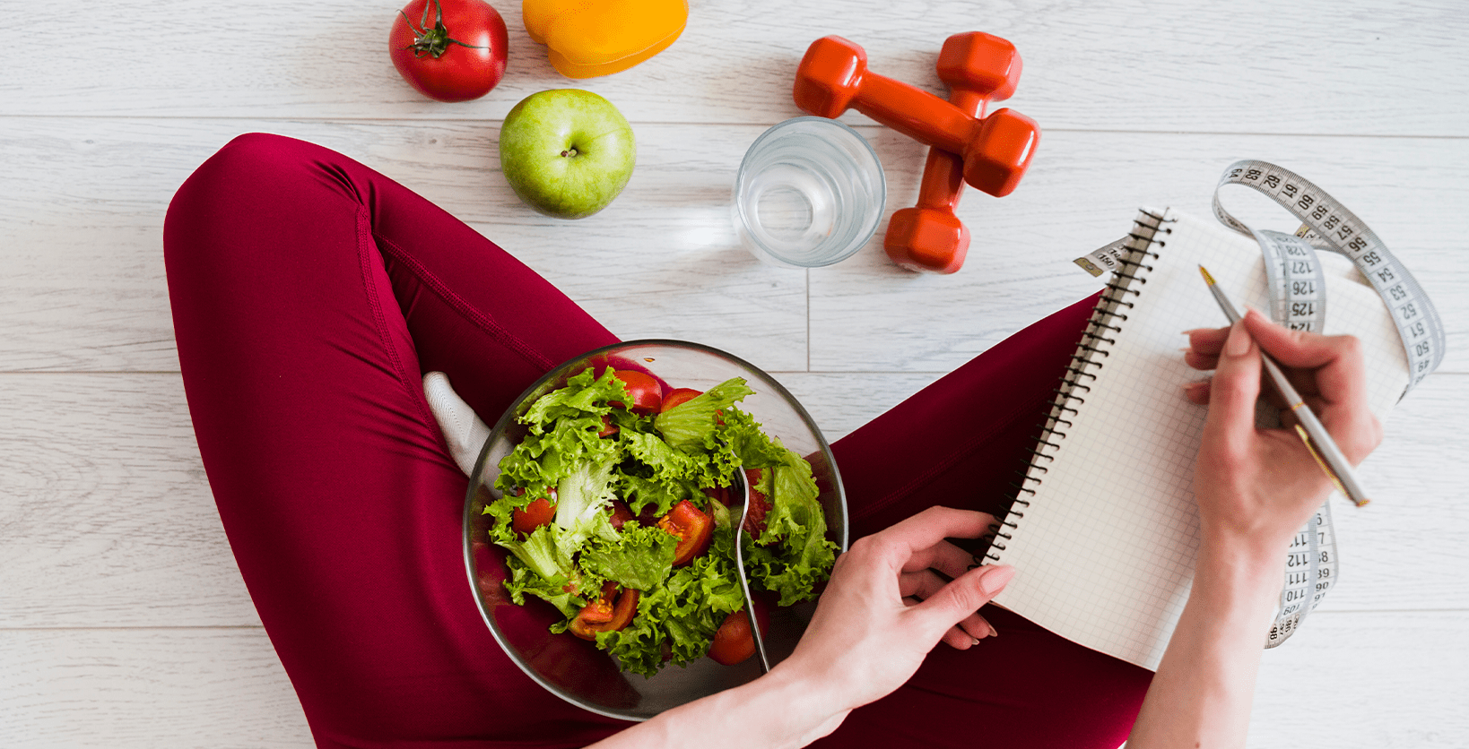 Woman sits on floor with bowl of salad on her lap and exercise weights on the floor in front of her.